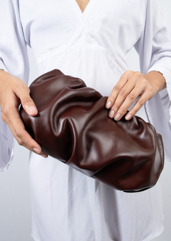 Brown leather clutch held by a person wearing a white outfit on a light gray background